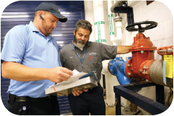 Photo: two men in polos (one blue, one gray) examine documents next to a backflow prevention device